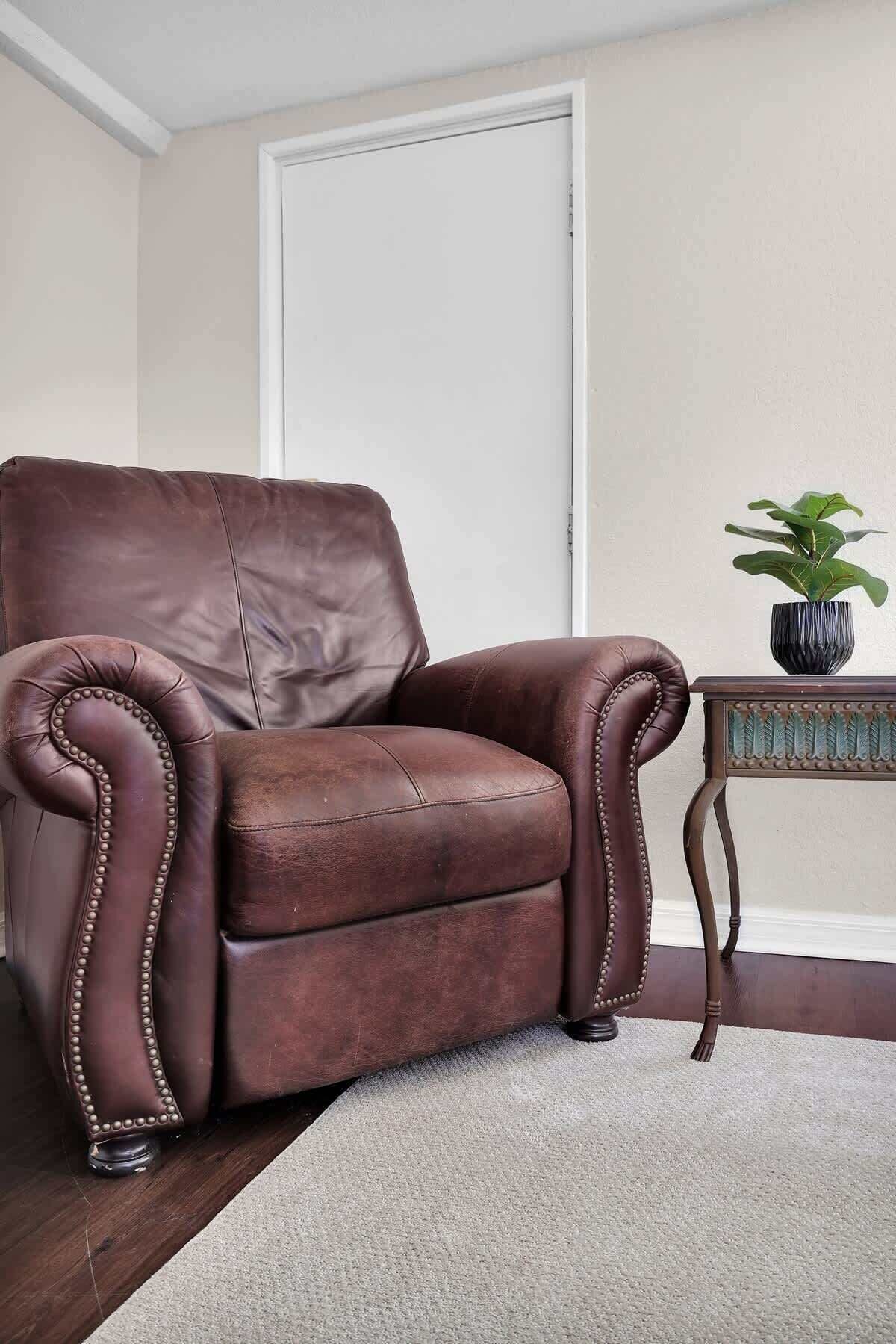 Un sillón de cuero marrón con respaldo alto y costuras decorativas, ubicado sobre un piso alfombrado junto a una pequeña mesa con una planta.