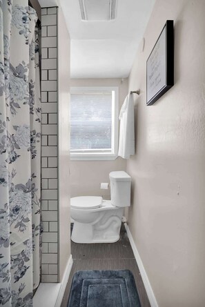 A bathroom with a toilet, a window with blinds, a shower curtain with a floral pattern, and a framed quote on the wall. - Smart Saver Townhouse, Family Friendly, Close to NAS Jax, Long-Term, Washer and Dryer (Jacksonville)