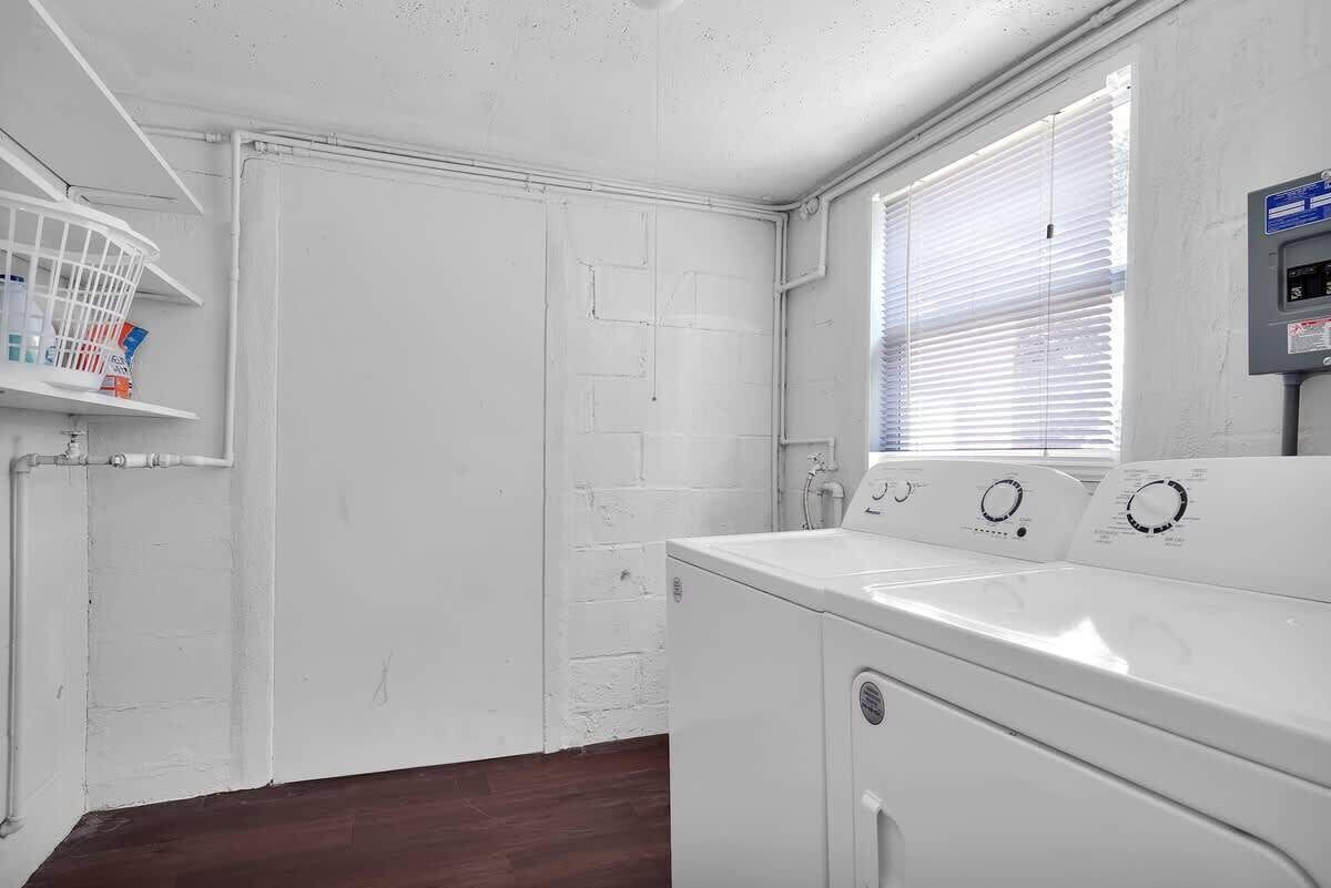 A laundry room with a white brick wall, a window with blinds, a washing machine, and a shelf with laundry supplies.