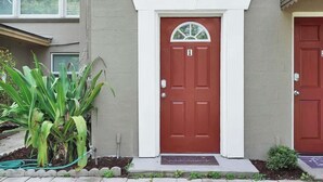 A two-story building with a red door, white trim, and a small garden area with plants. - Smart Saver Townhouse, Family Friendly, Close to NAS Jax, Long-Term, Washer and Dryer (Jacksonville)
