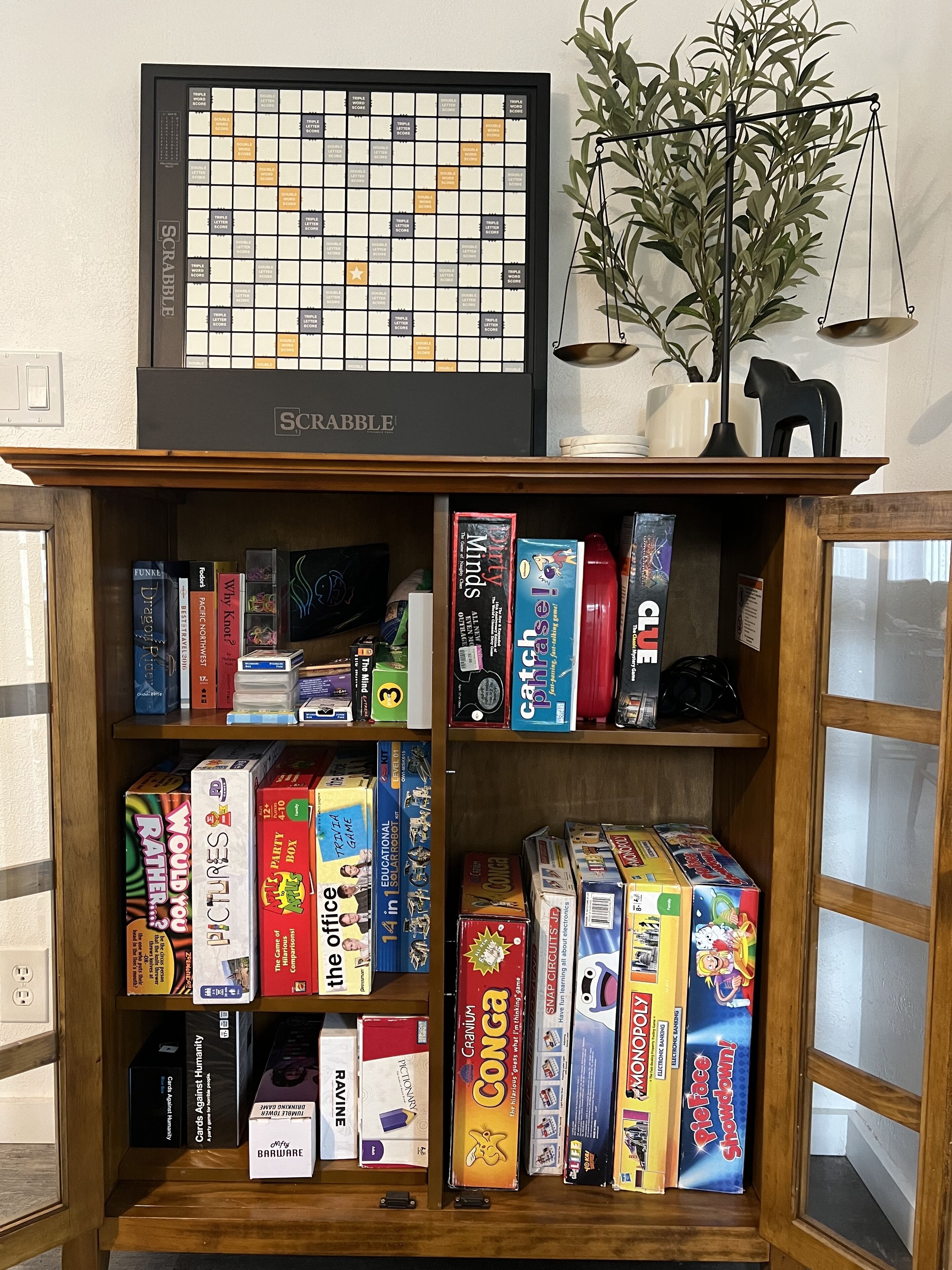 A Scrabble board game on a shelf above a wooden cabinet filled with various board games.
