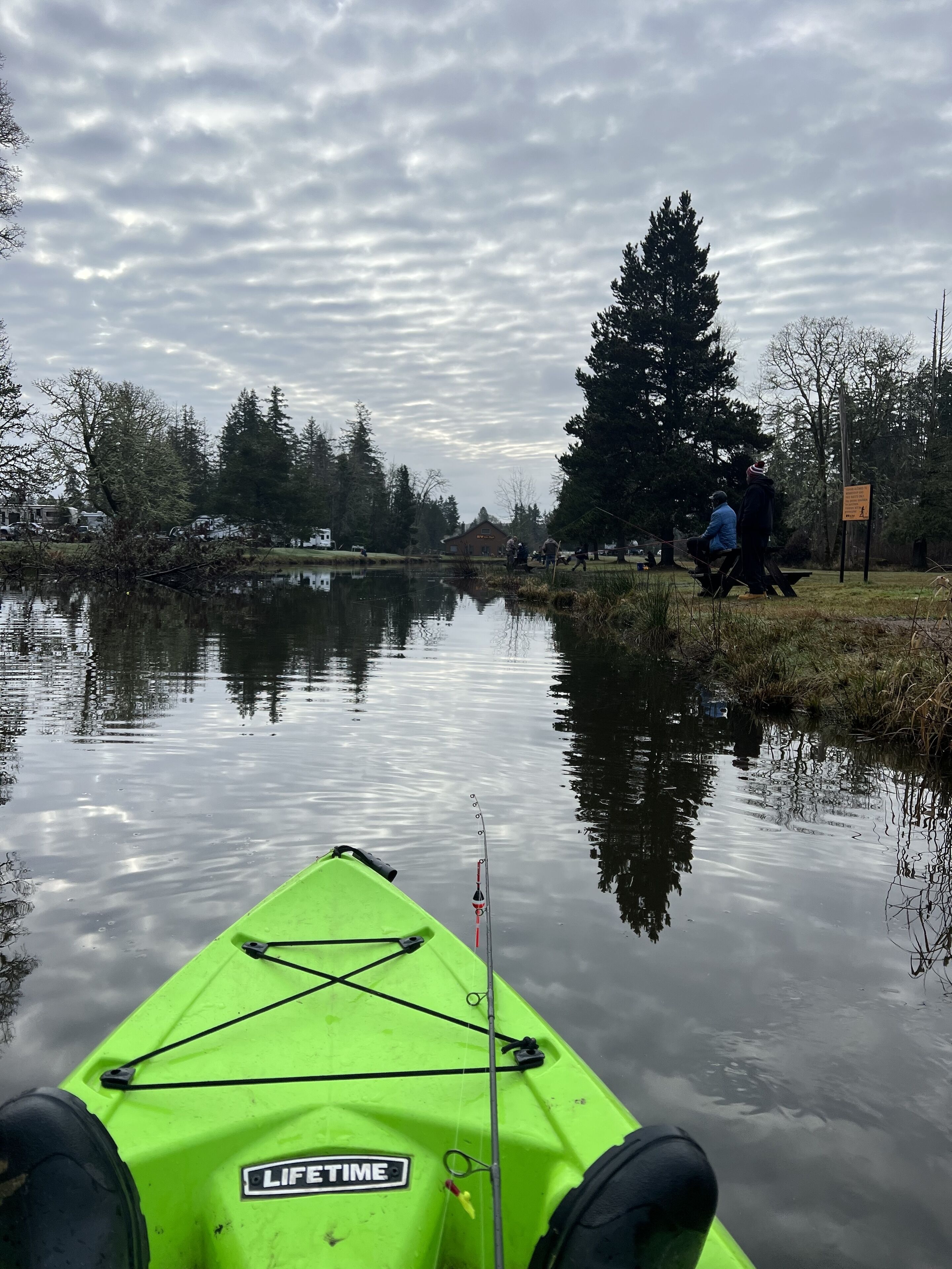 Un kayak verde con una caña de pescar, un río tranquilo y un área de picnic con gente al fondo.