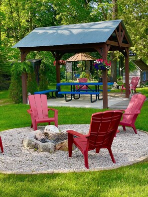 A gazebo with a picnic table and red chairs, surrounded by a fire pit and gravel.