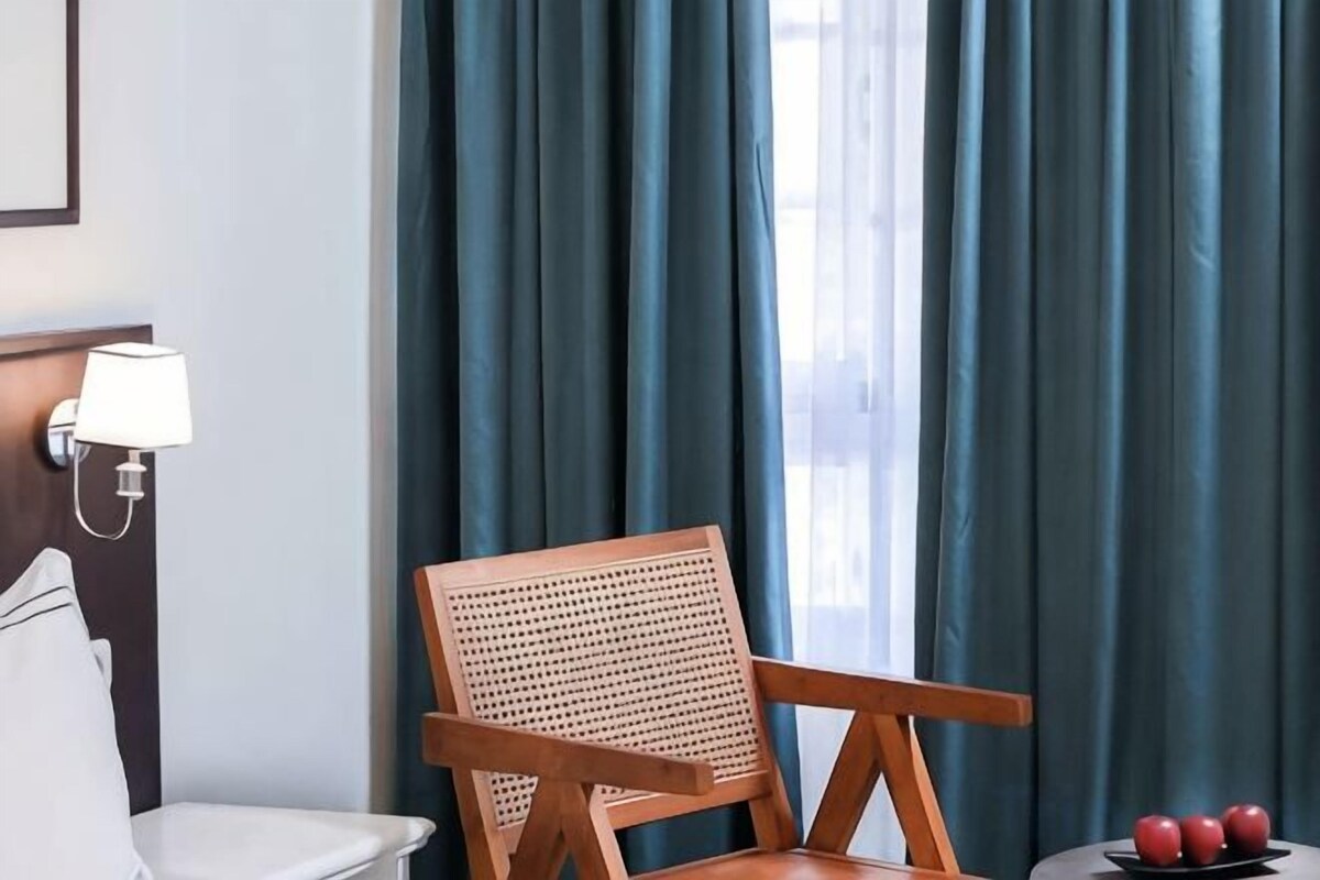 A hotel room with a wooden chair, a small table with apples, and a window with curtains.