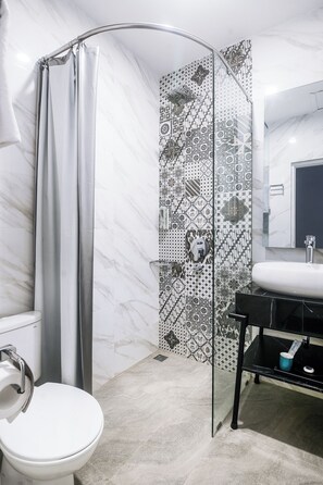 A bathroom with a white toilet, a shower with a glass door, a sink with a black countertop, and patterned tiles on the walls. - Hotel Noola Malioboro (Yogyakarta)