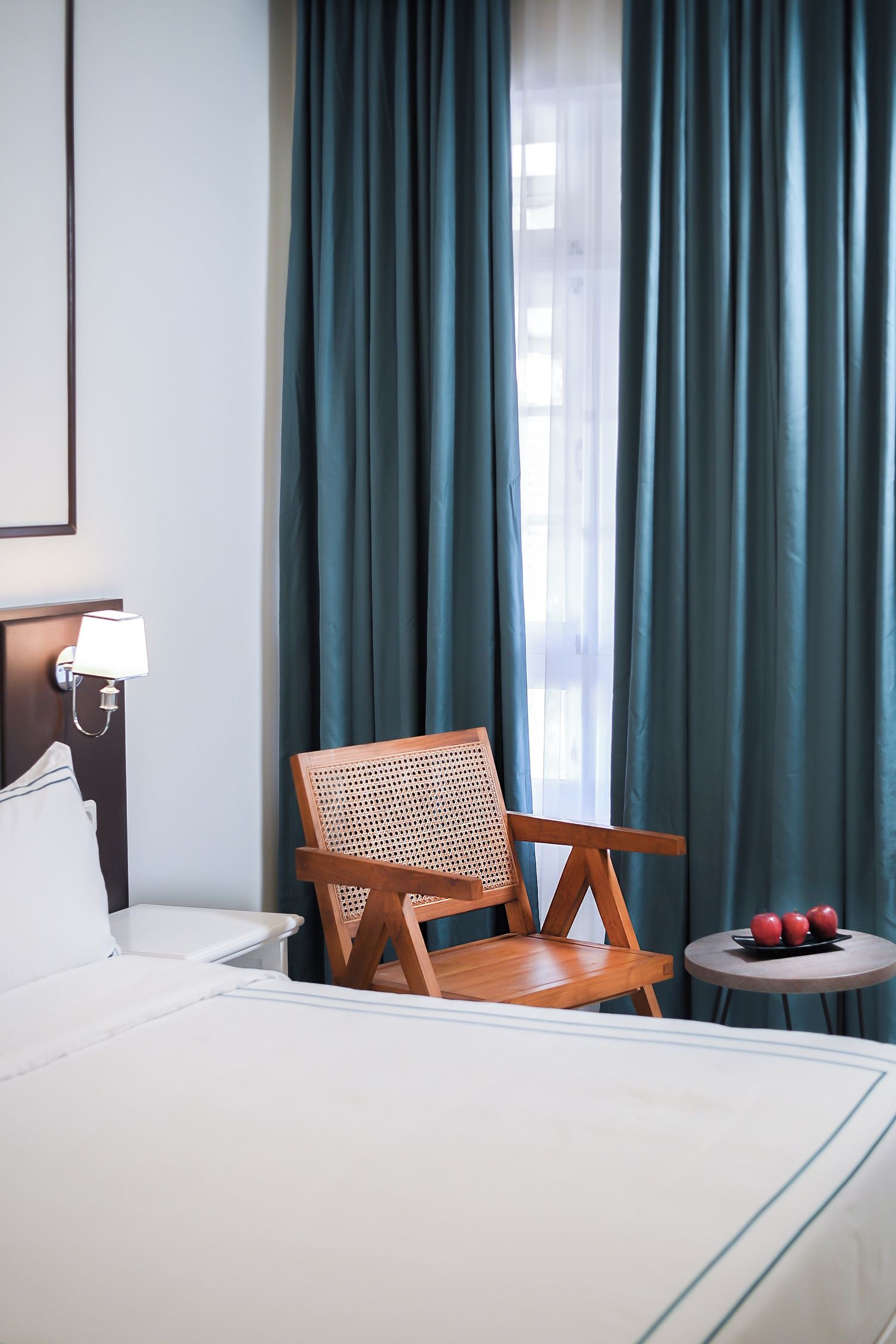 A hotel room with a bed, a wooden chair with a woven backrest, a small round table with apples, and a lamp.