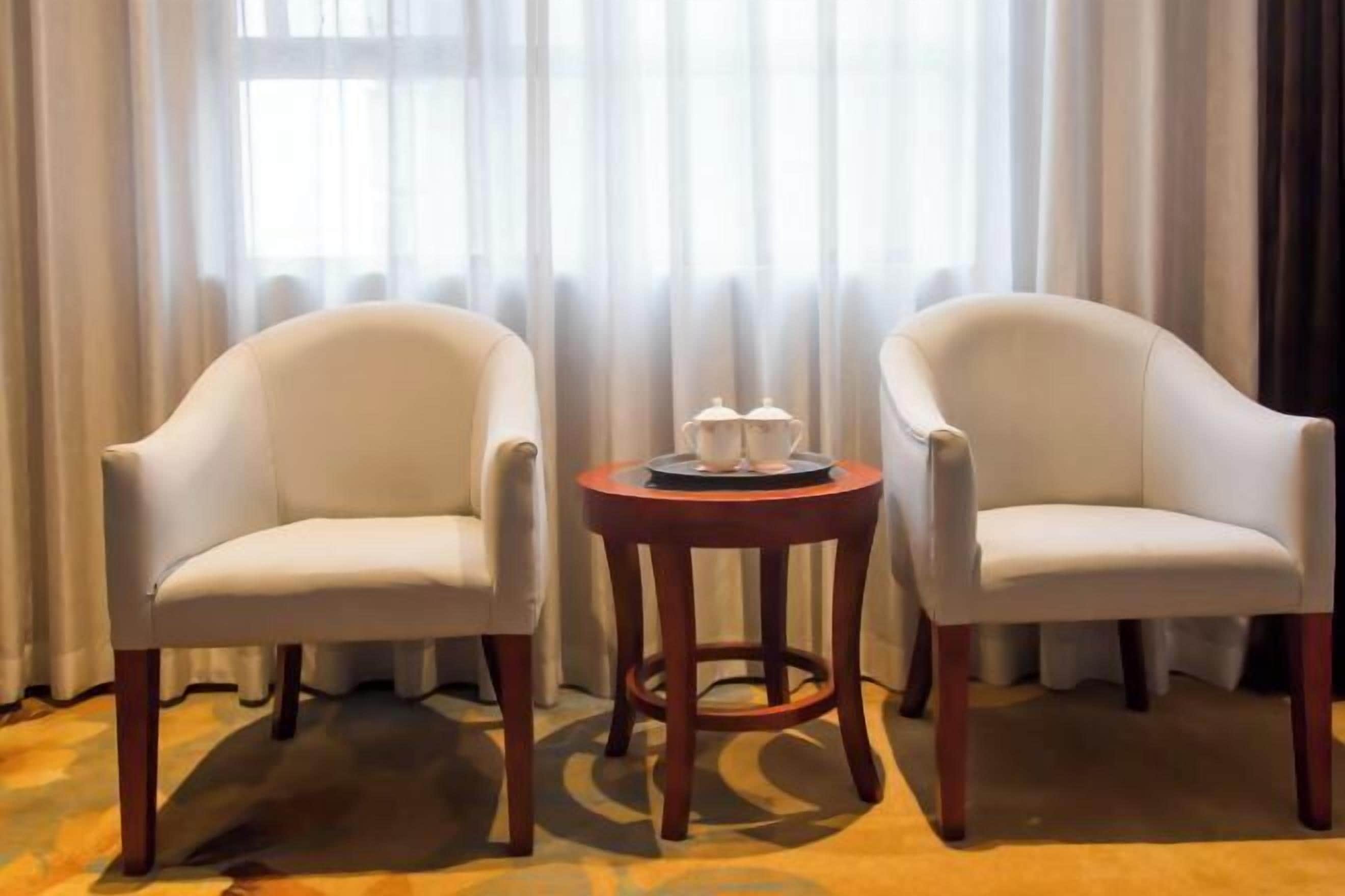Dos sillones blancos y una pequeña mesa de madera con una bandeja de tazas y platillos en una habitación con un ventanal y cortinas translúcidas.