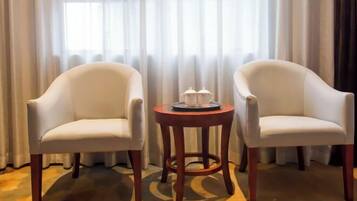 Dos sillones blancos y una pequeña mesa de madera con una bandeja de tazas y platillos en una habitación con un ventanal y cortinas translúcidas.