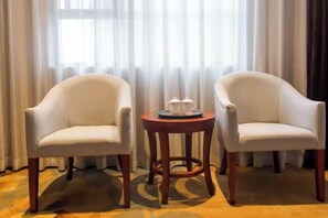 Dos sillones blancos y una pequeña mesa de madera con una bandeja de tazas y platillos en una habitación con un ventanal y cortinas translúcidas.