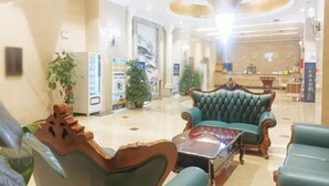 A lobby with a reception desk, two green upholstered armchairs, a coffee table, and potted plants. - Tongxilai Hotel (Jingyan)