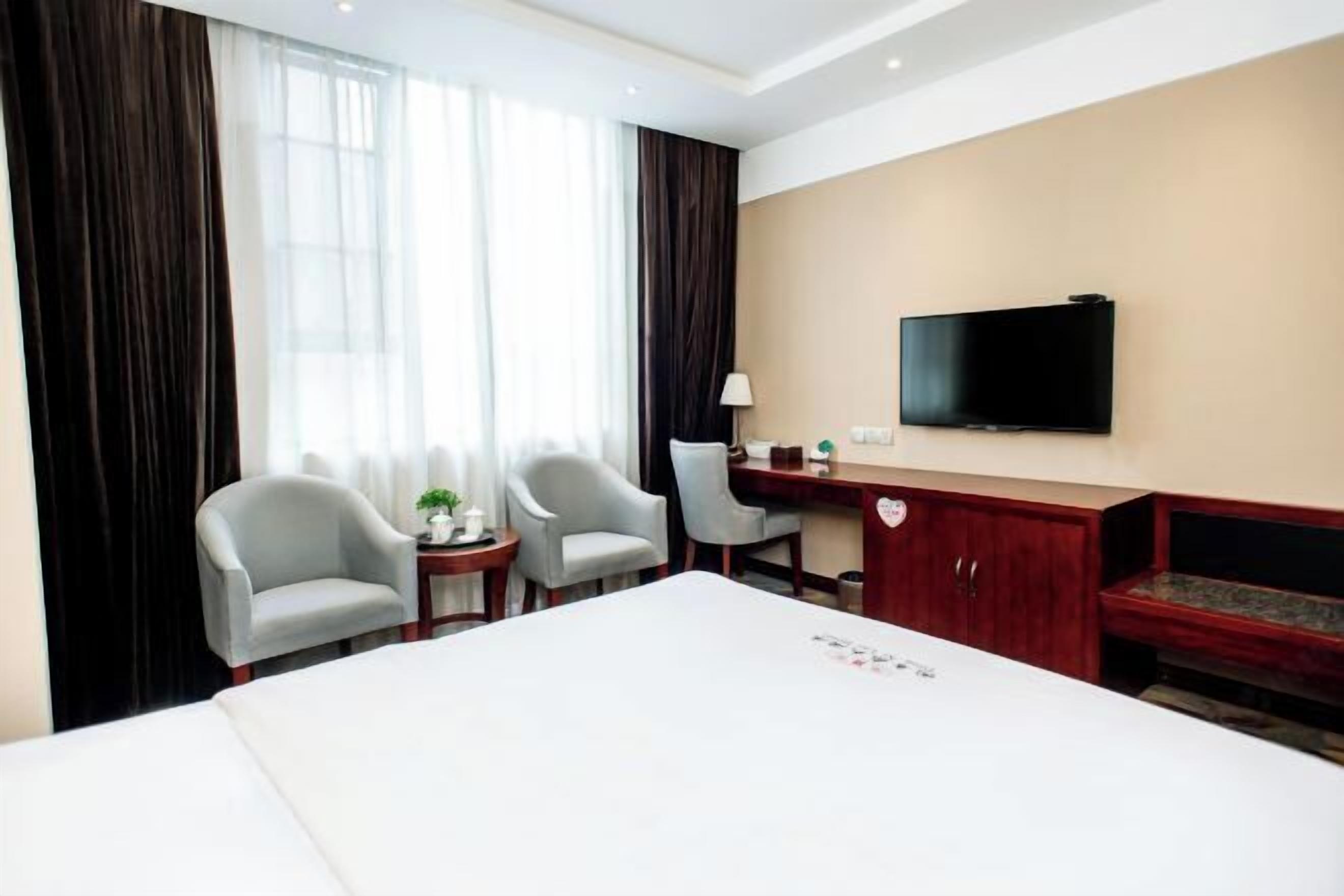 Une chambre d’hôtel avec un grand lit, deux fauteuils, un bureau avec une télévision et une fenêtre avec des rideaux.