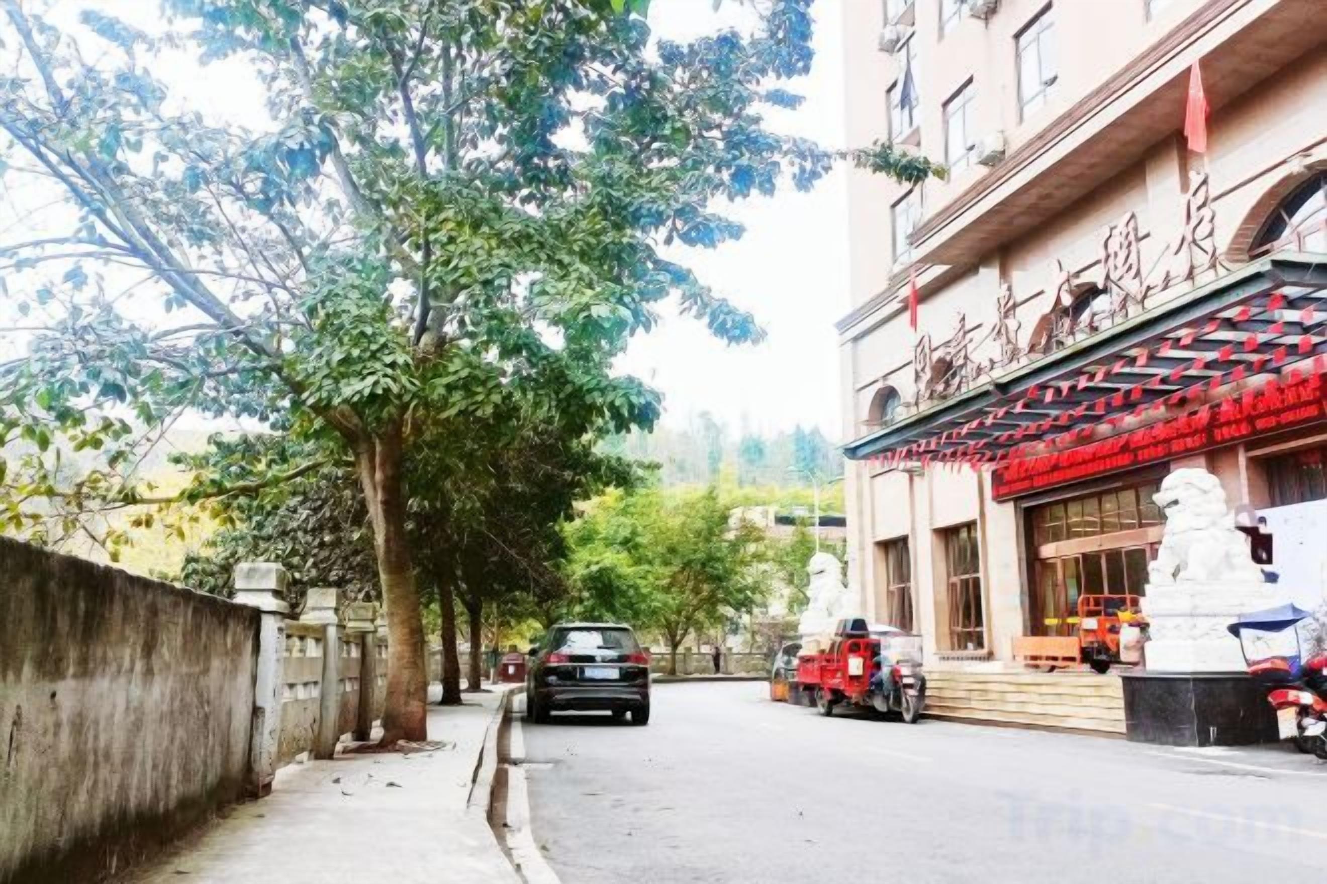 A street view with a building featuring a traditional architectural style, a tree-lined sidewalk, and parked vehicles.