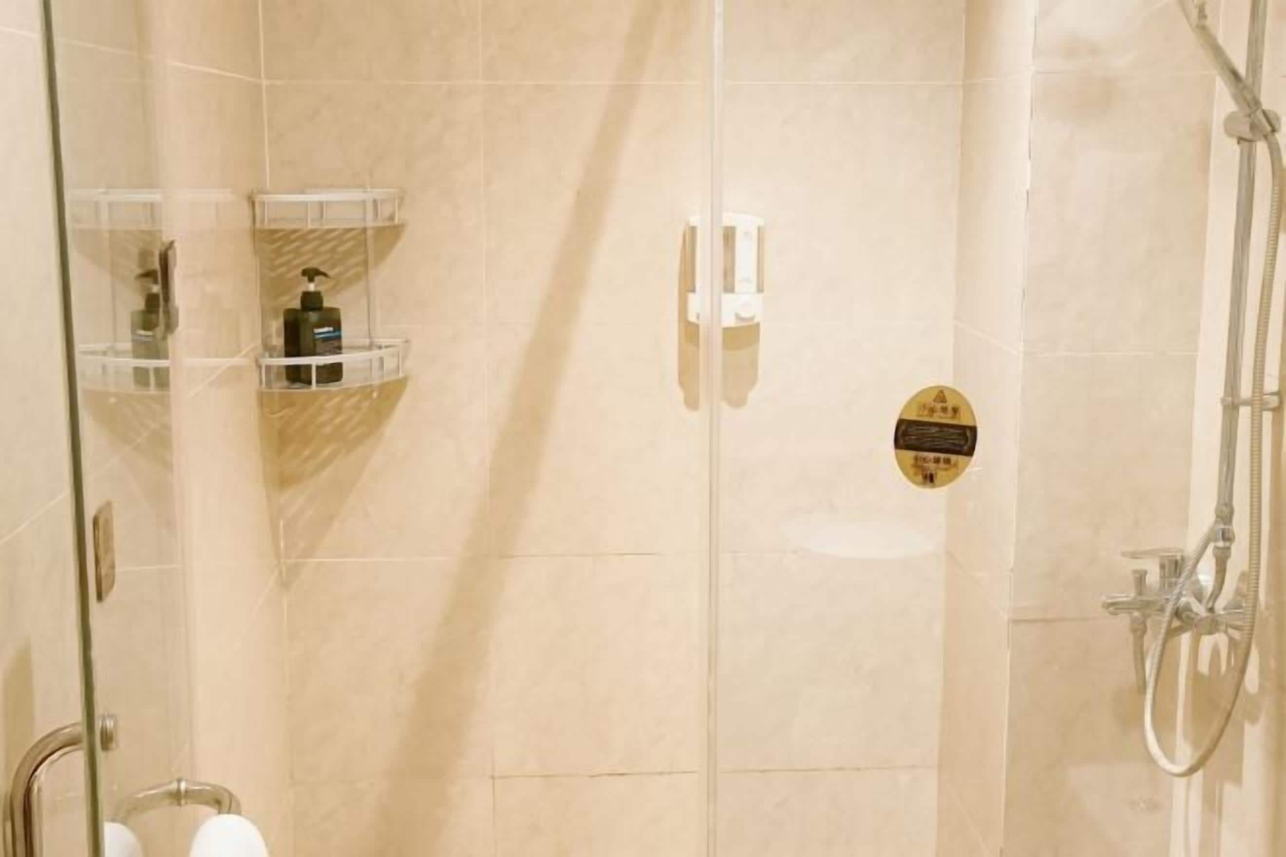 A glass-enclosed shower area with a handheld showerhead, a glass shelf with toiletries, and a safety sign on the wall.
