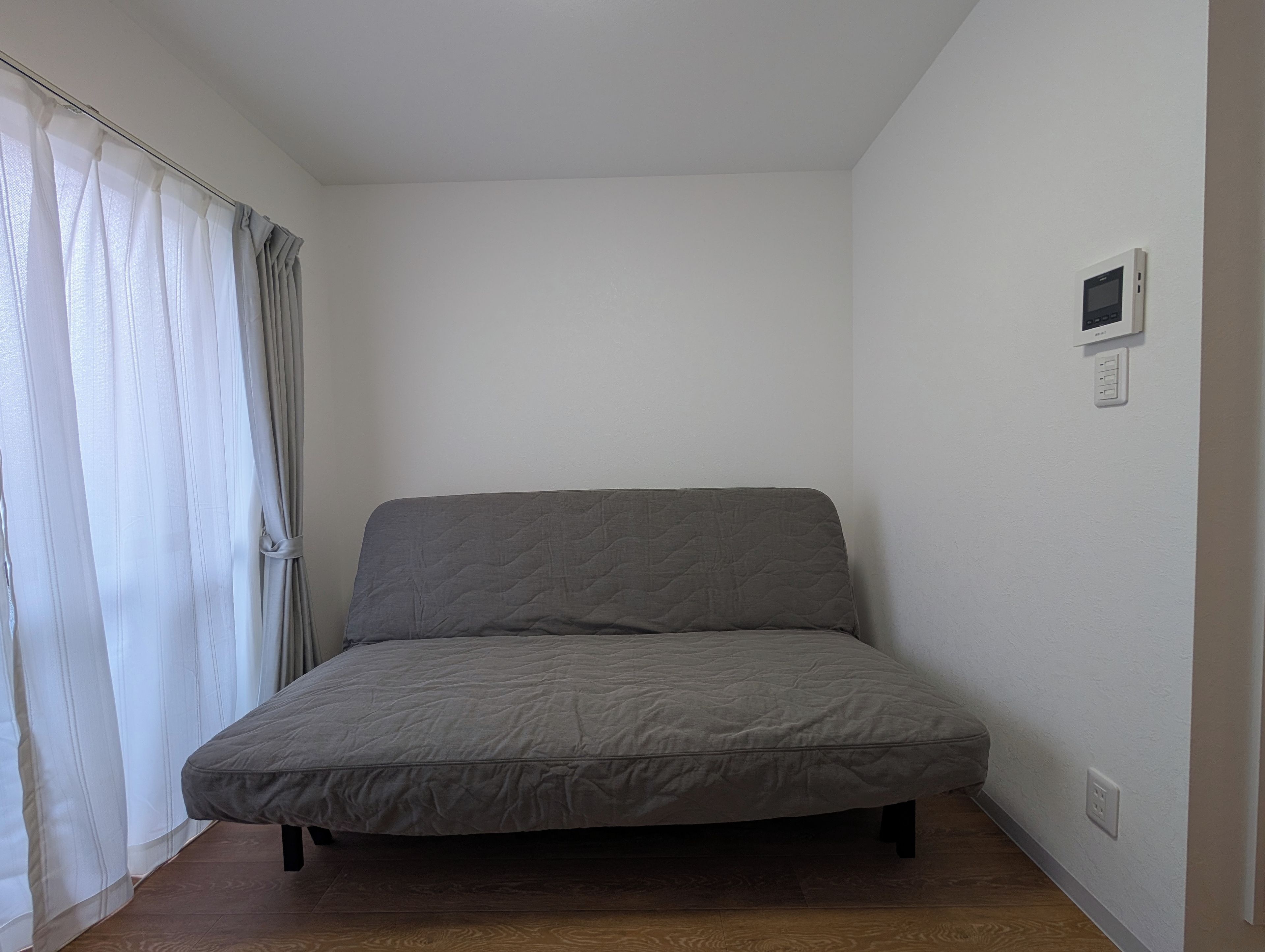 A single bed in a room with a wooden floor, white walls, and a window with white curtains.