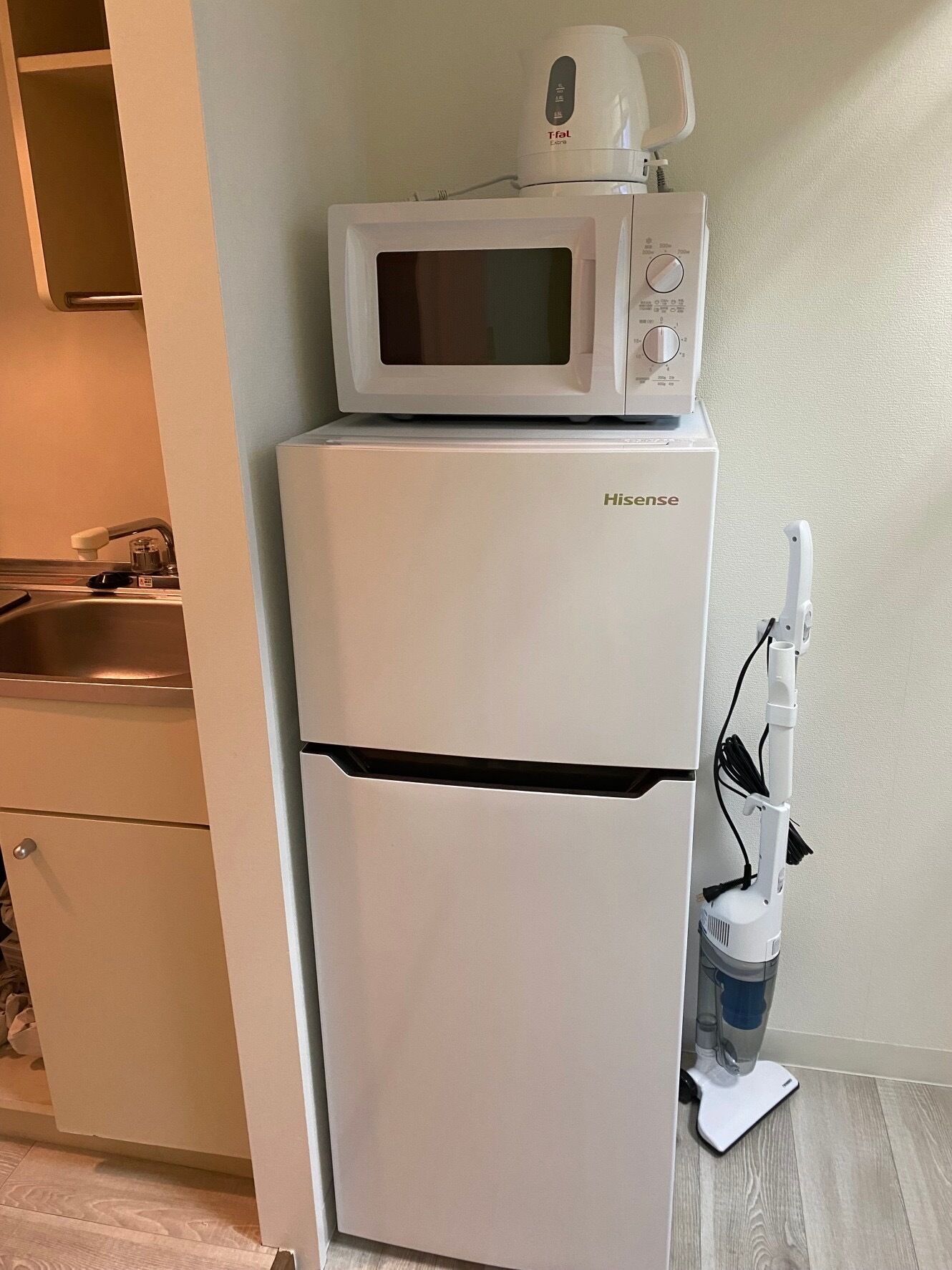 A compact kitchenette with a white refrigerator, a microwave, and a vacuum cleaner.