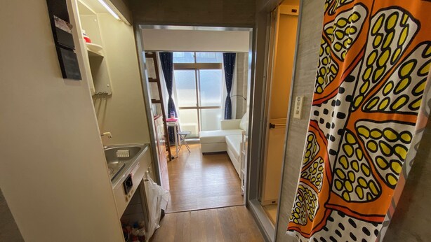A narrow hallway with a kitchen area on the left, a white sofa in the center, and a patterned curtain on the right. - Quartier Blanc Yoga (Tokyo)