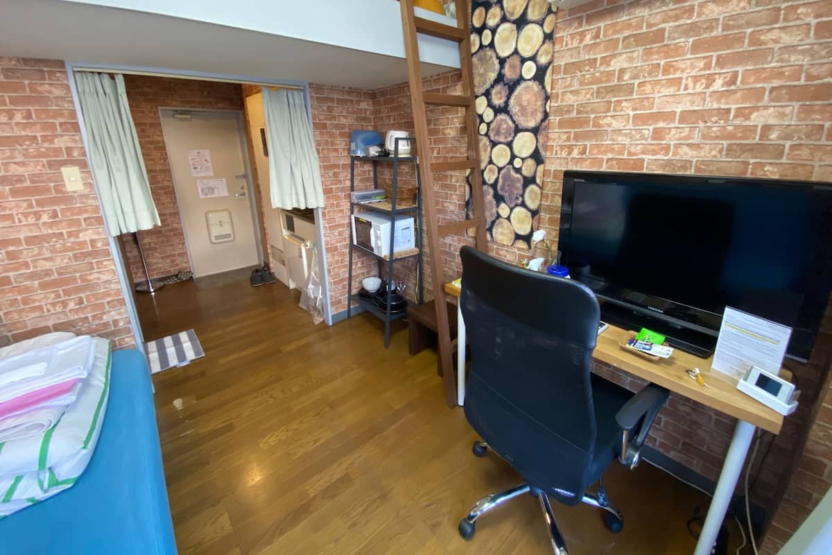 A room with a brick wall, wooden flooring, a desk with a computer, a TV, and a bed with a blue cover.