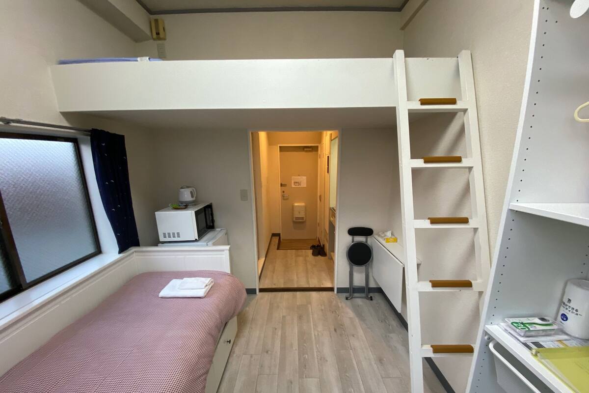 A compact room with a bunk bed, a desk with a computer, a microwave, and a small kitchenette.
