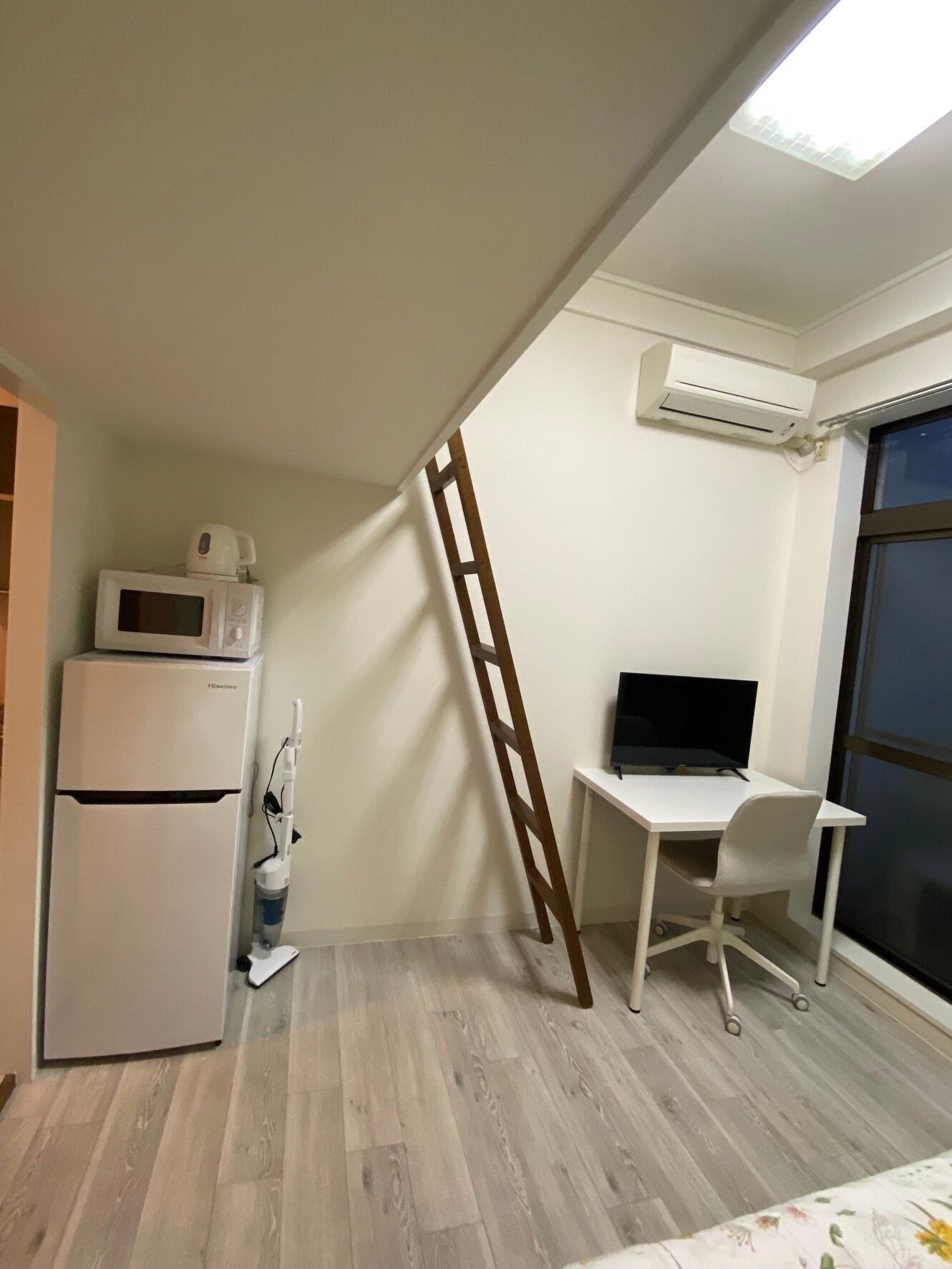 A compact room with a kitchenette, a desk with a computer, a ladder, and a large window.