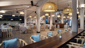 A restaurant with high ceilings, exposed beams, and a long wooden bar. Tables are set with white tablecloths and blue cushions. - The Urban  (Siem Reap)