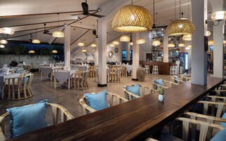Restaurante com tetos altos, vigas aparentes e um longo balcão de madeira. Mesas com toalhas brancas e almofadas azuis.