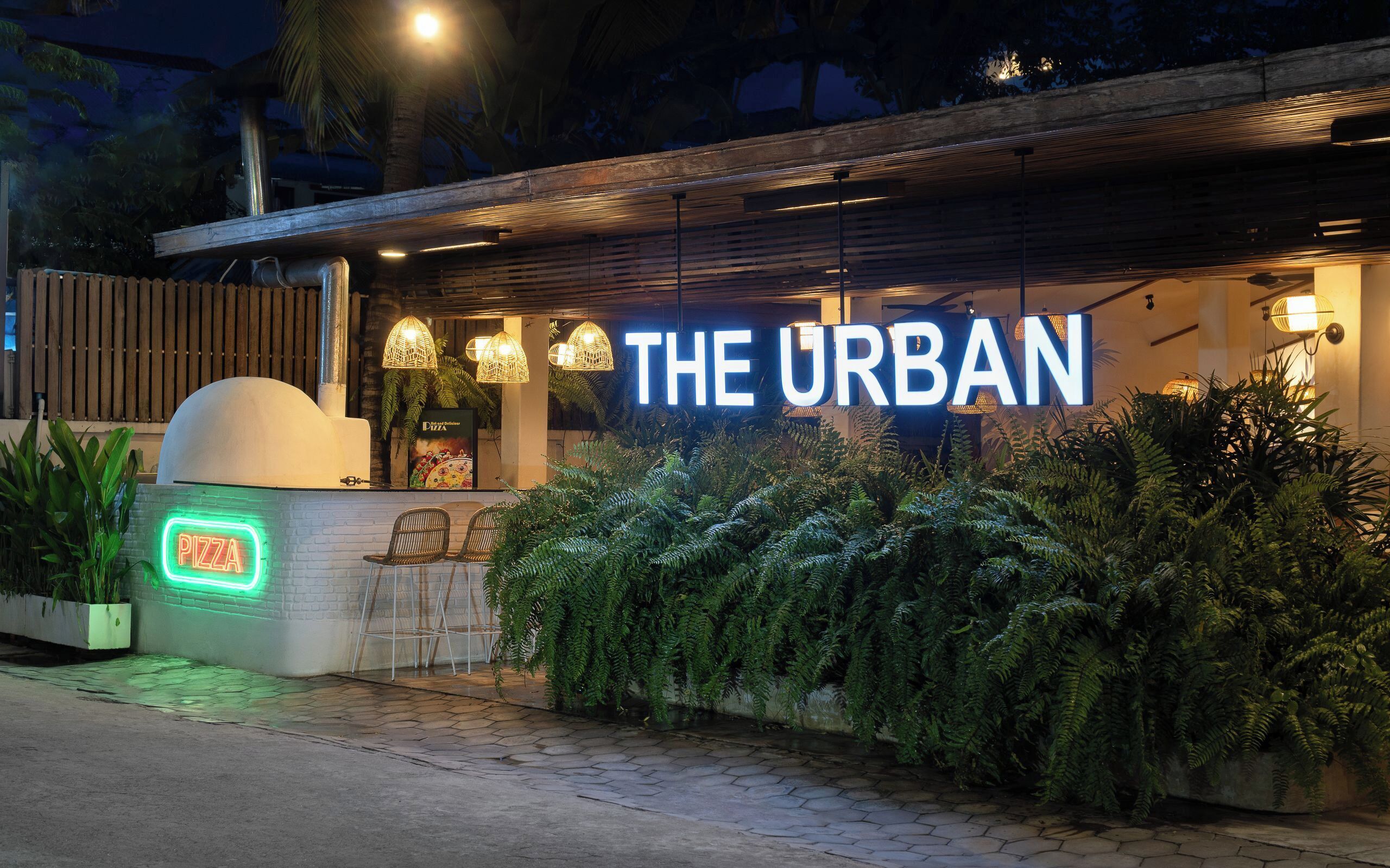 Un restaurante llamado "The Urban" con un letrero de neón, mesas al aire libre y abundante vegetación.