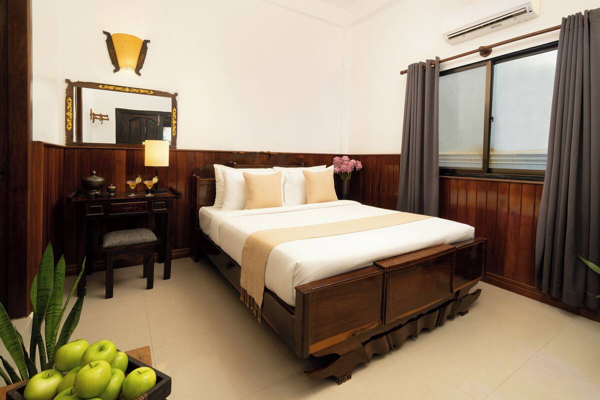 Una habitación de hotel con una cama de madera, un escritorio con lámpara, un espejo y una ventana con cortinas.