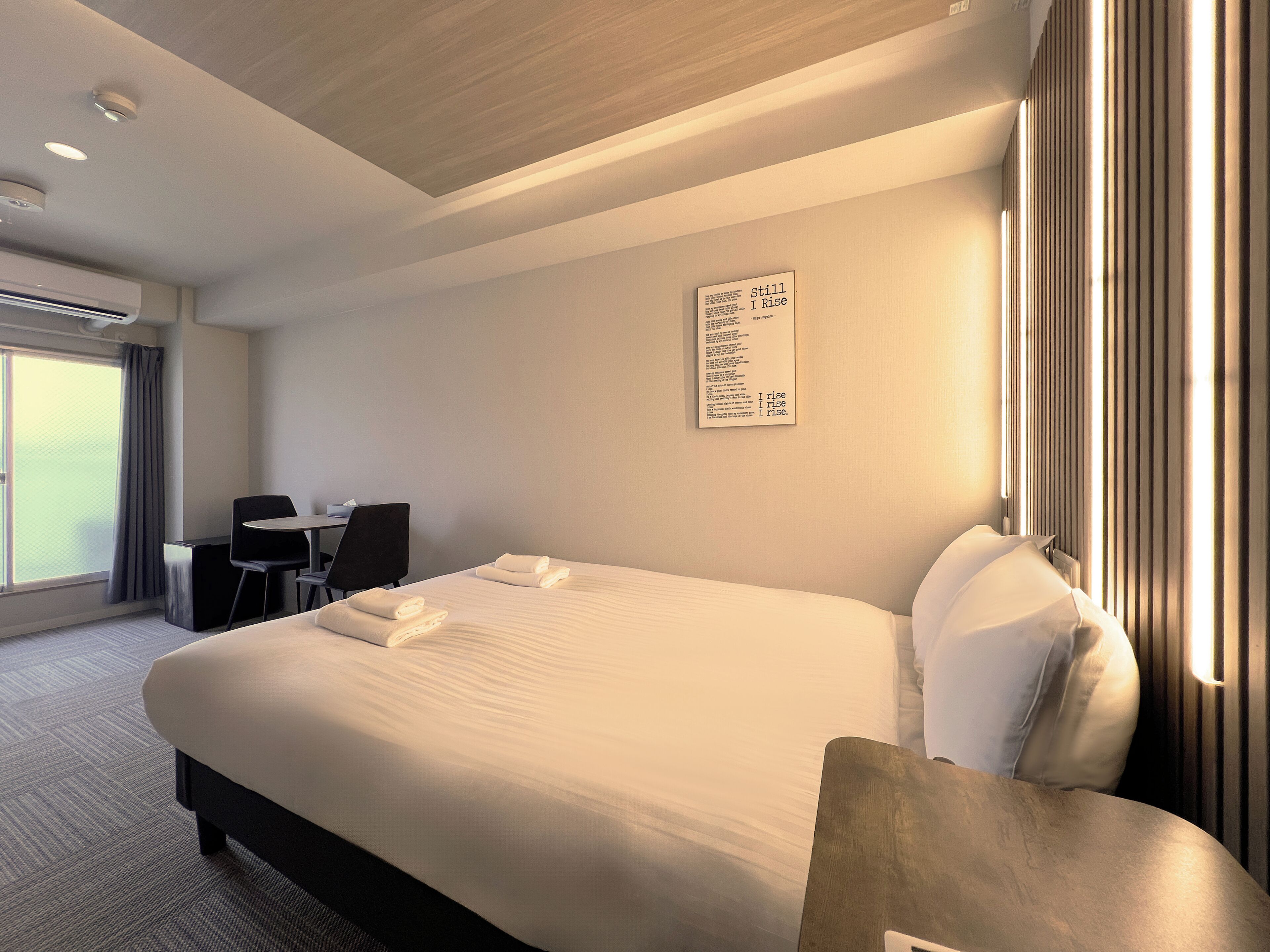 A hotel room with a large bed, a desk, and a chair. There is a window with curtains and a wall-mounted artwork.