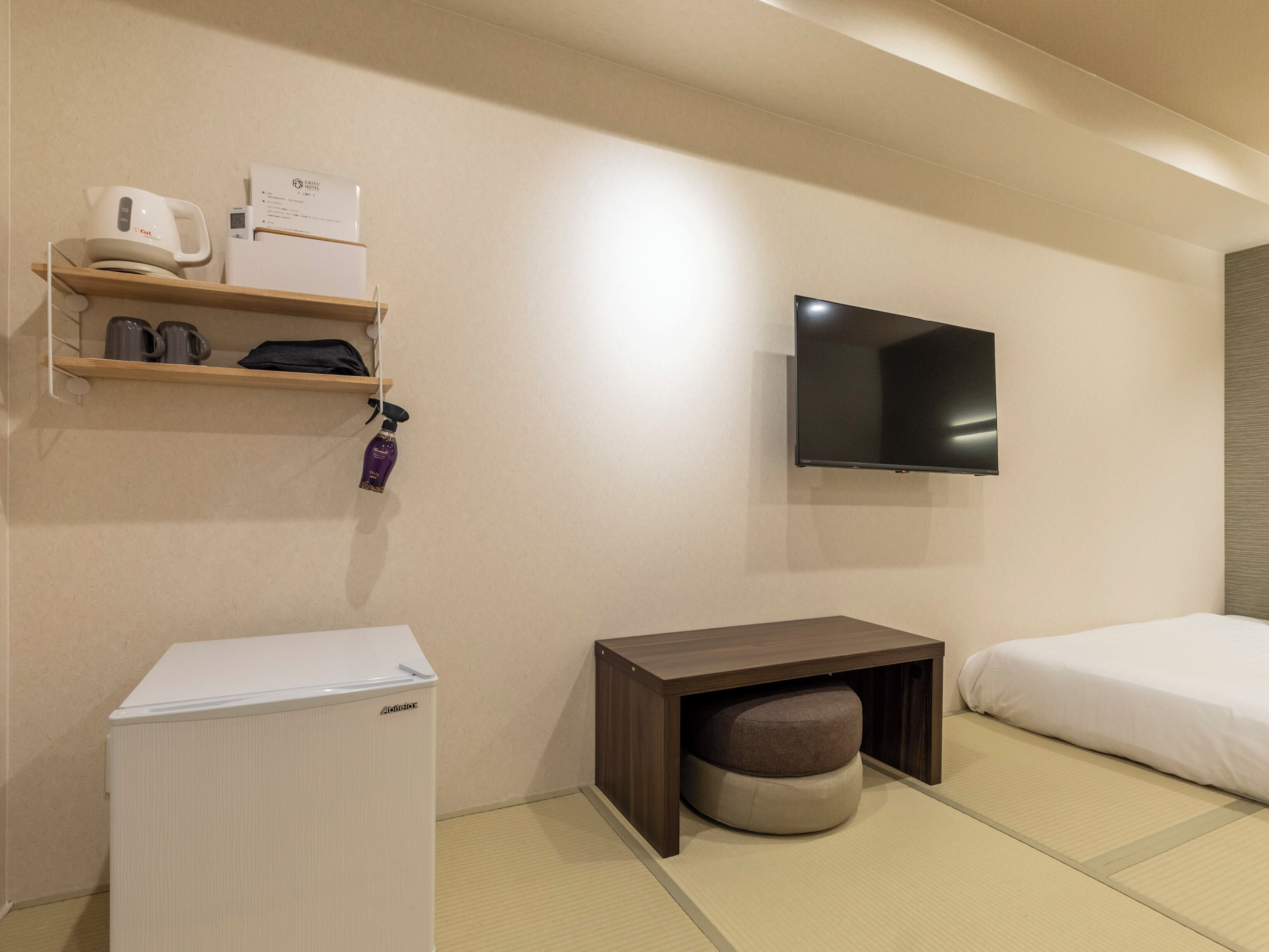 A hotel room with a bed, a TV mounted on the wall, a small wooden table, a white refrigerator, and a shelf with toiletries.