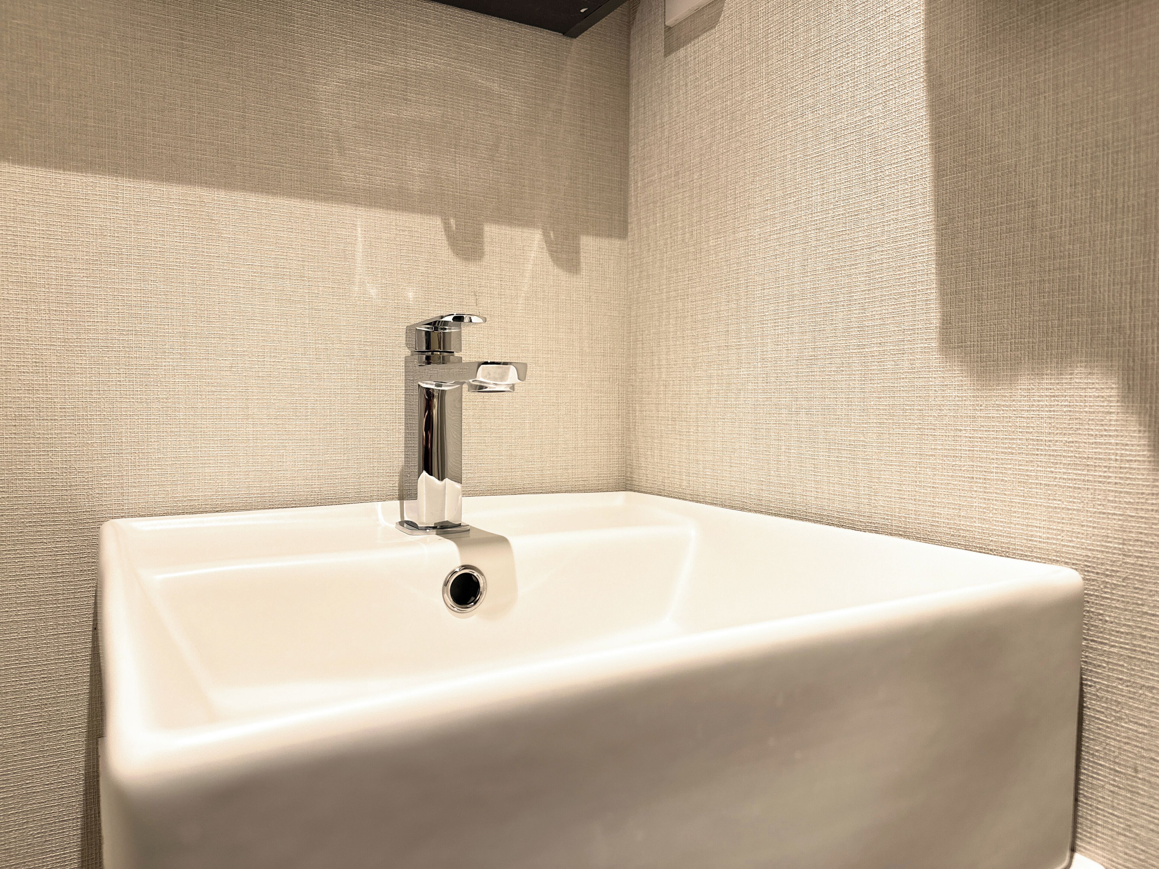 A white bathroom sink with a chrome faucet against a textured beige wall.