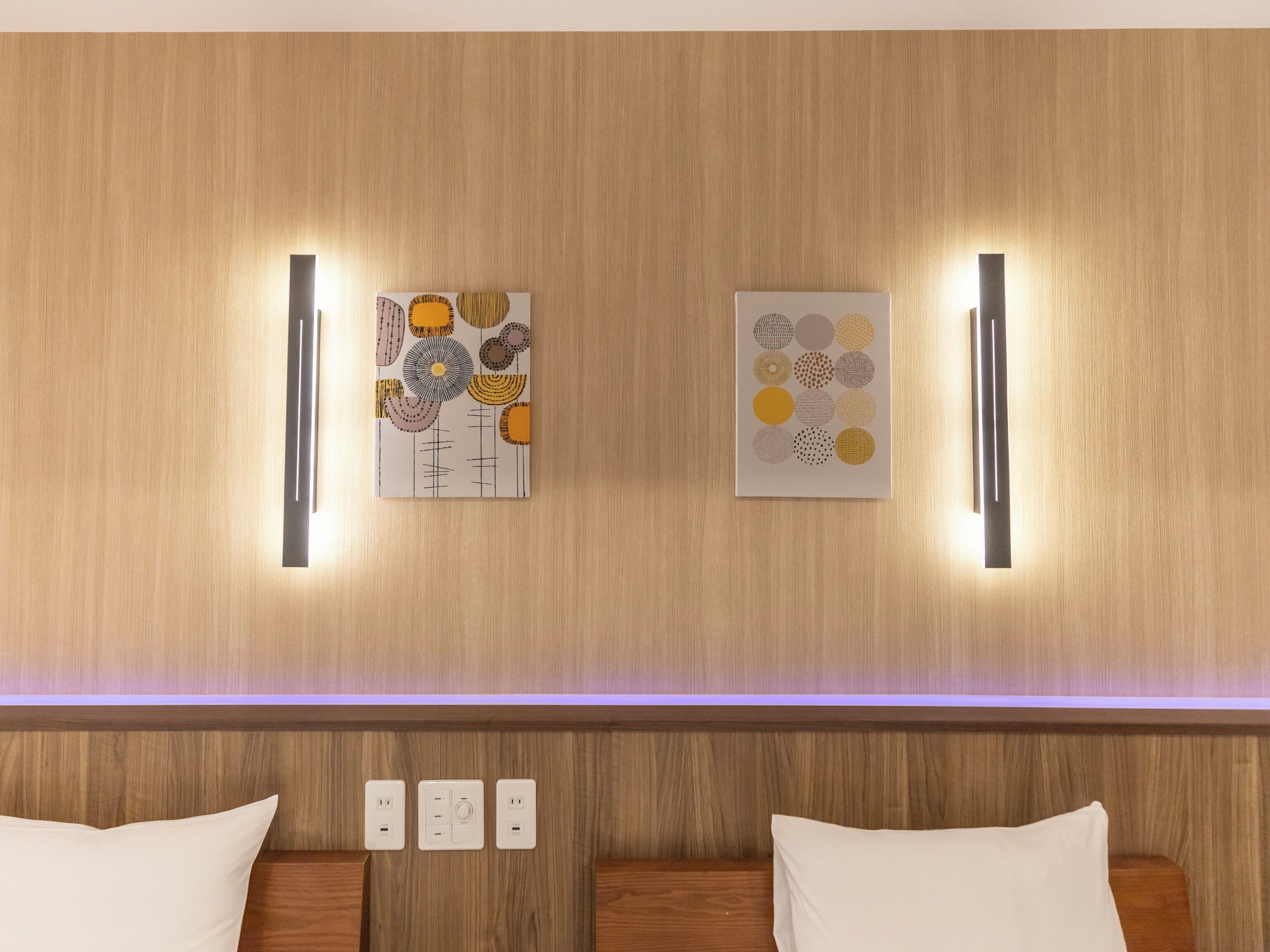 A hotel room with a wooden wall, two artworks, a bedside table, and a bed with white bedding.