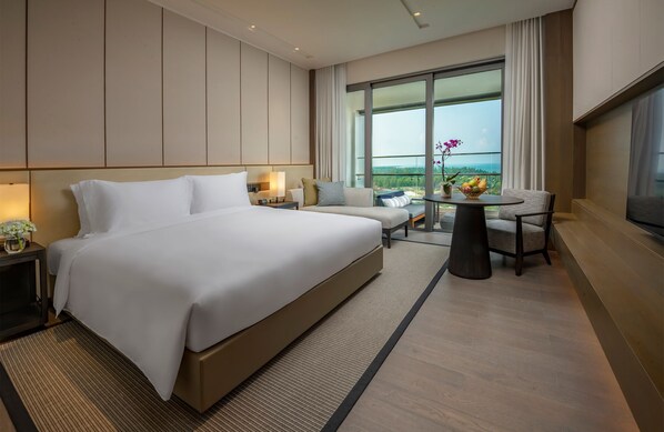 A modern hotel room with a large bed, a sofa, a dining table with fruit, and a view of the outdoors.
