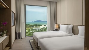 A hotel room with two beds, a large window with curtains, a bedside table with a lamp, and a view of the sea and mountains. - Hoiana Residences (Da Nang)
