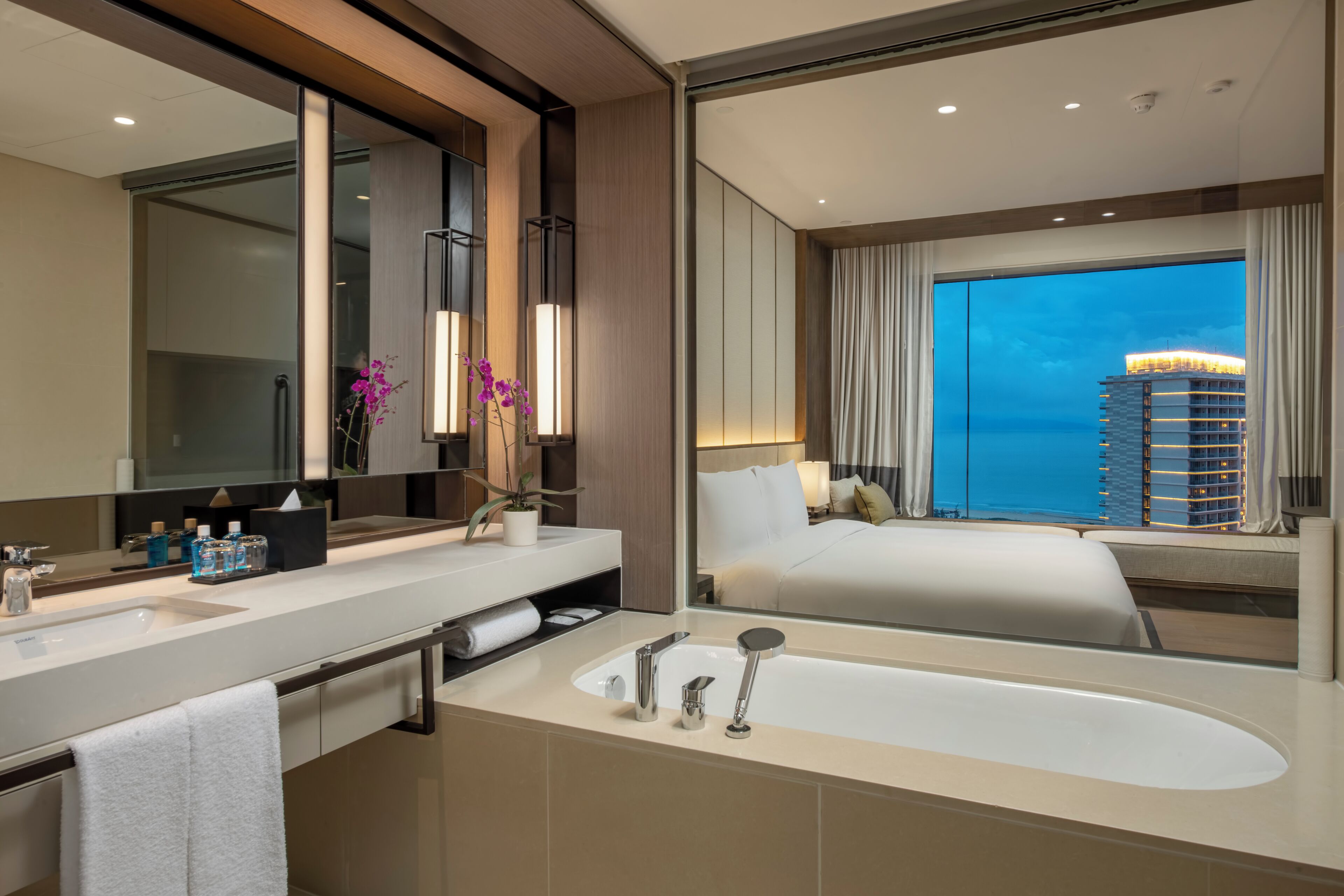 Ein modernes Hotelbad mit einer großen Badewanne, einem Schminktisch mit Spiegel und Blick auf eine Stadtlandschaft durch ein großes Fenster.