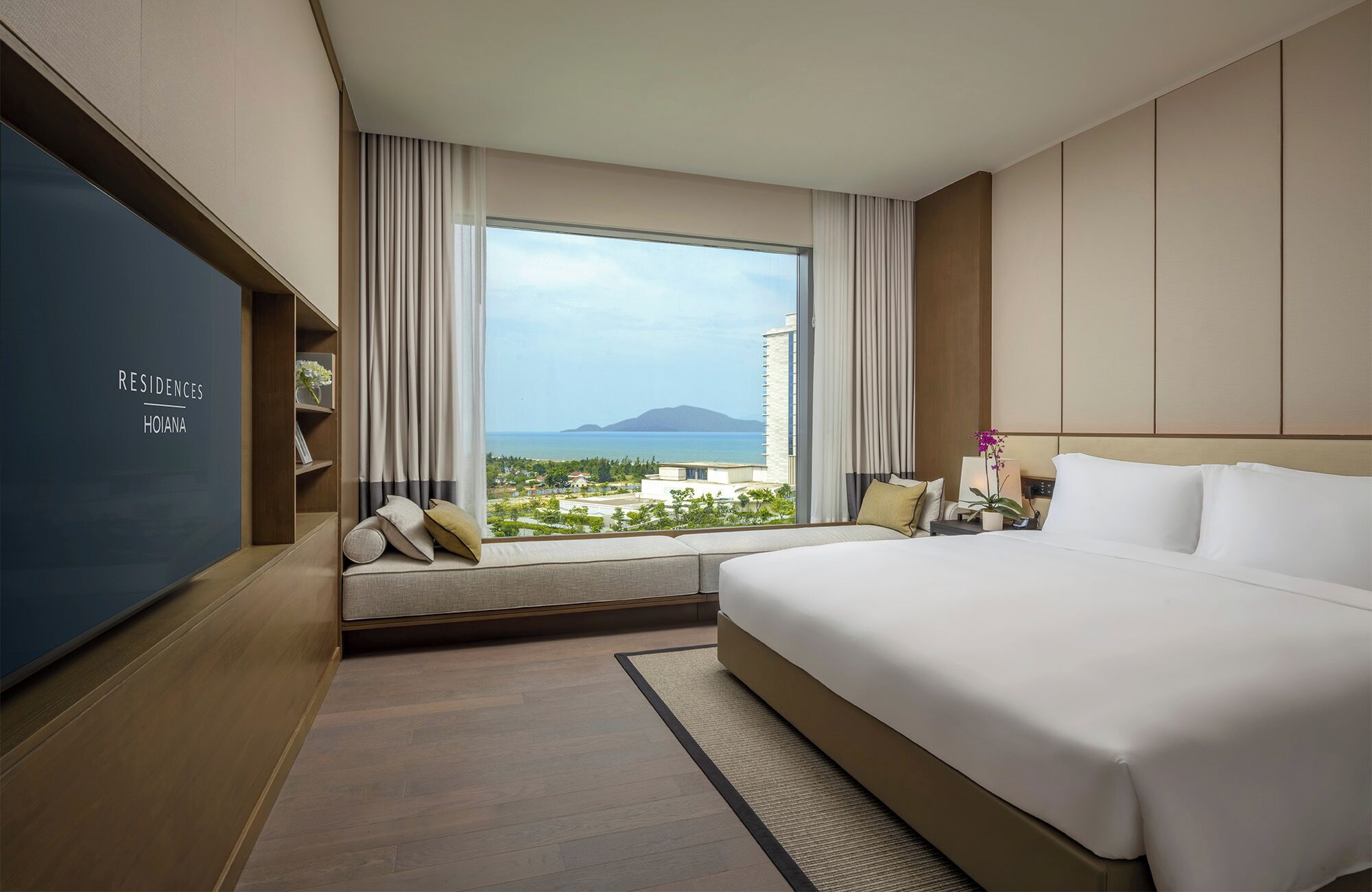 A hotel room with a large bed, a TV, a window with a view of the sea and buildings, and a small seating area.
