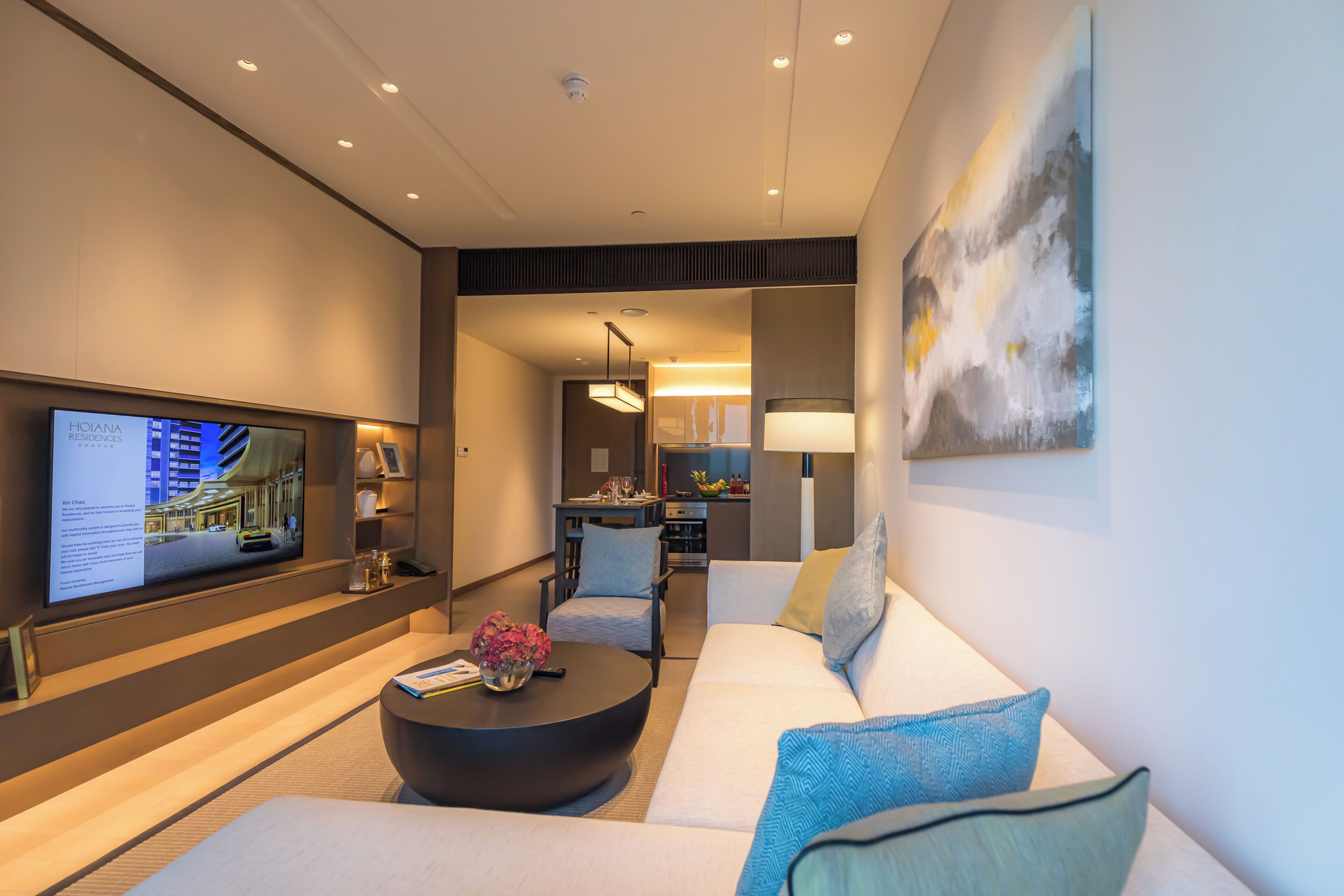 A modern hotel room with a flat-screen TV, a round coffee table, a sofa, and a dining area with a lamp.