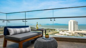A balcony with a sofa, a round ottoman, a small table with a bottle and glass, and a view of the sea and buildings. - Hoiana Residences (Da Nang)