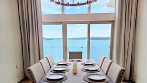 A dining area with a table set for six, large windows with a view of the sea, and a chandelier. - *NEW* TV-Featured Mansion / Waterfront / Dock / Golf Green / Games (Sunrise Beach)