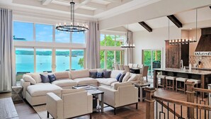 A spacious living room with a large L-shaped sectional sofa, a wooden dining table with bar stools, and a view of the water through large windows. - *NEW* TV-Featured Mansion / Waterfront / Dock / Golf Green / Games (Sunrise Beach)