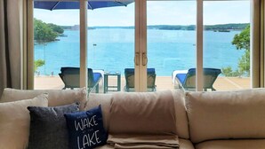 A room with a large window overlooking a lake, a beige sofa with a pillow, and two chairs outside. - *NEW* TV-Featured Mansion / Waterfront / Dock / Golf Green / Games (Sunrise Beach)