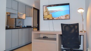 A modern kitchen with grey cabinets, a built-in oven, and a sink. There is a desk with a chair and a lamp, and a TV mounted on the wall displaying a scenic image. - Le Mansion Senopati Hotel (Jakarta)