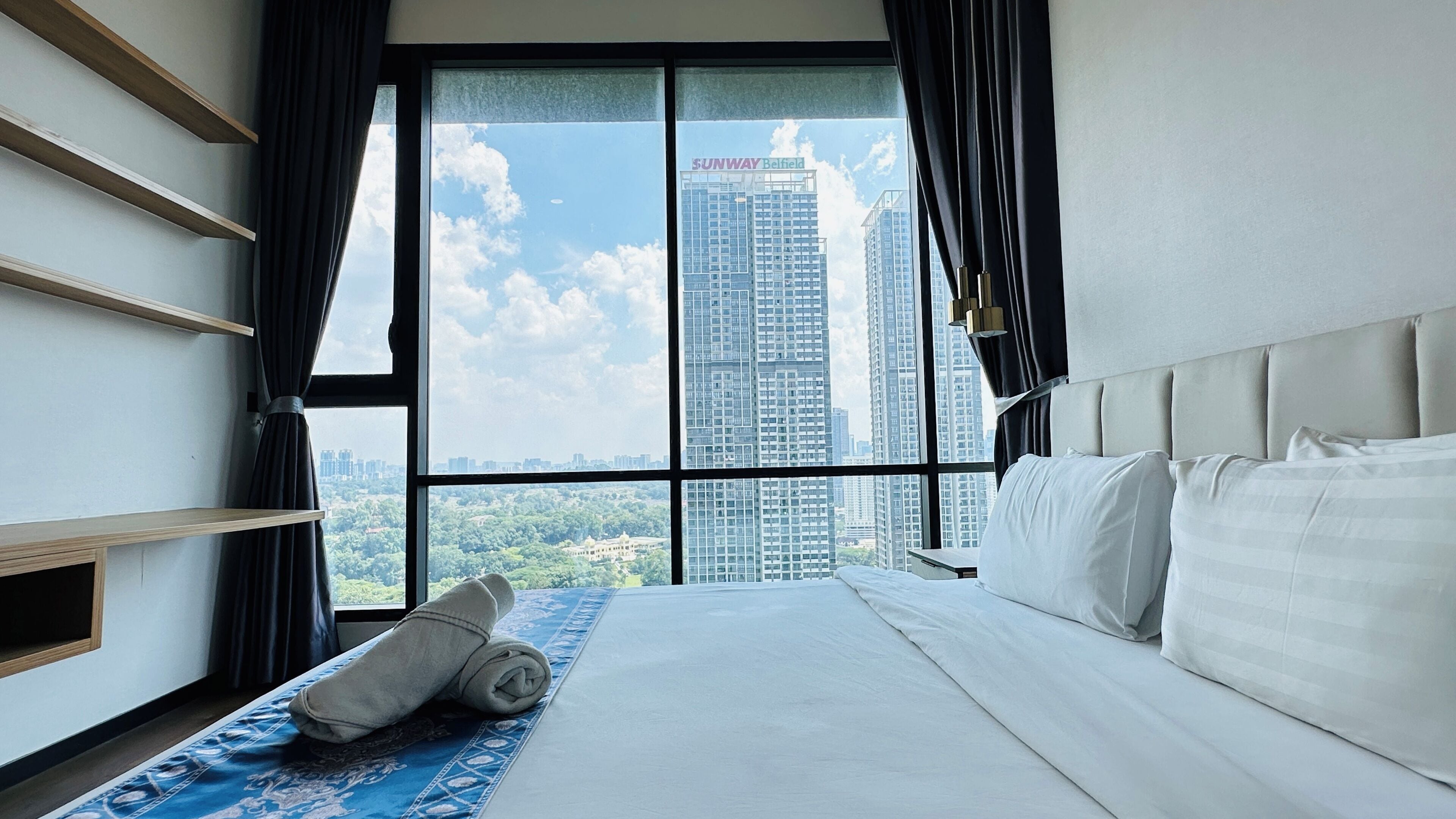 A hotel room with a large window, a bed, a blue patterned rug, and a view of high-rise buildings.