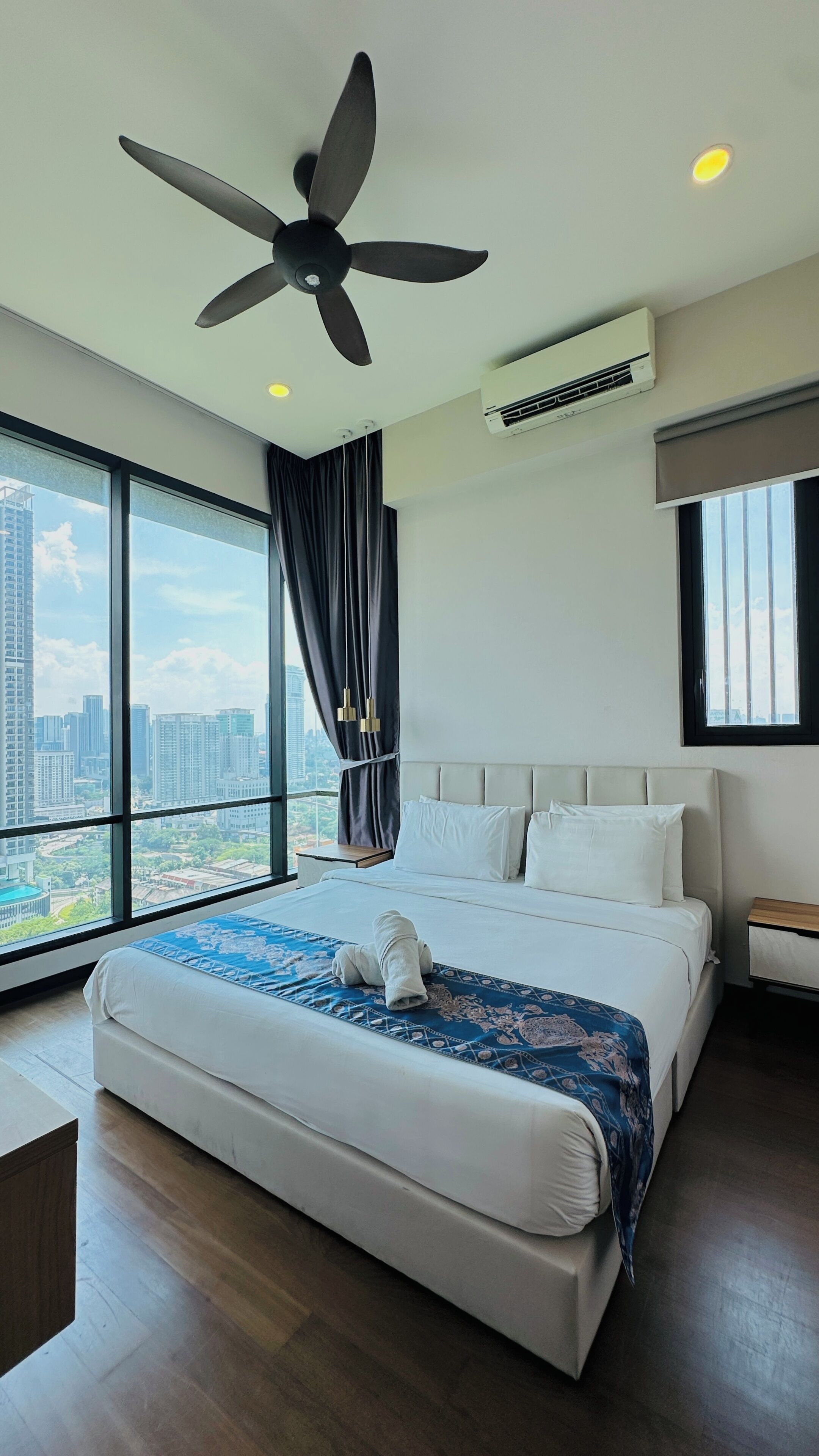 Una habitación de hotel moderna con una cama grande, ventilador de techo, unidad de aire acondicionado, vista a la ciudad y un ventanal con cortinas.
