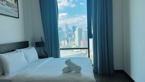 Quarto de hotel com cama, mesa de cabeceira, luminária e vista para a cidade através de grandes janelas.