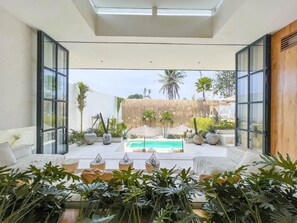 A modern indoor pool area with a glass ceiling, surrounded by tropical plants and furnished with white sofas and tables.