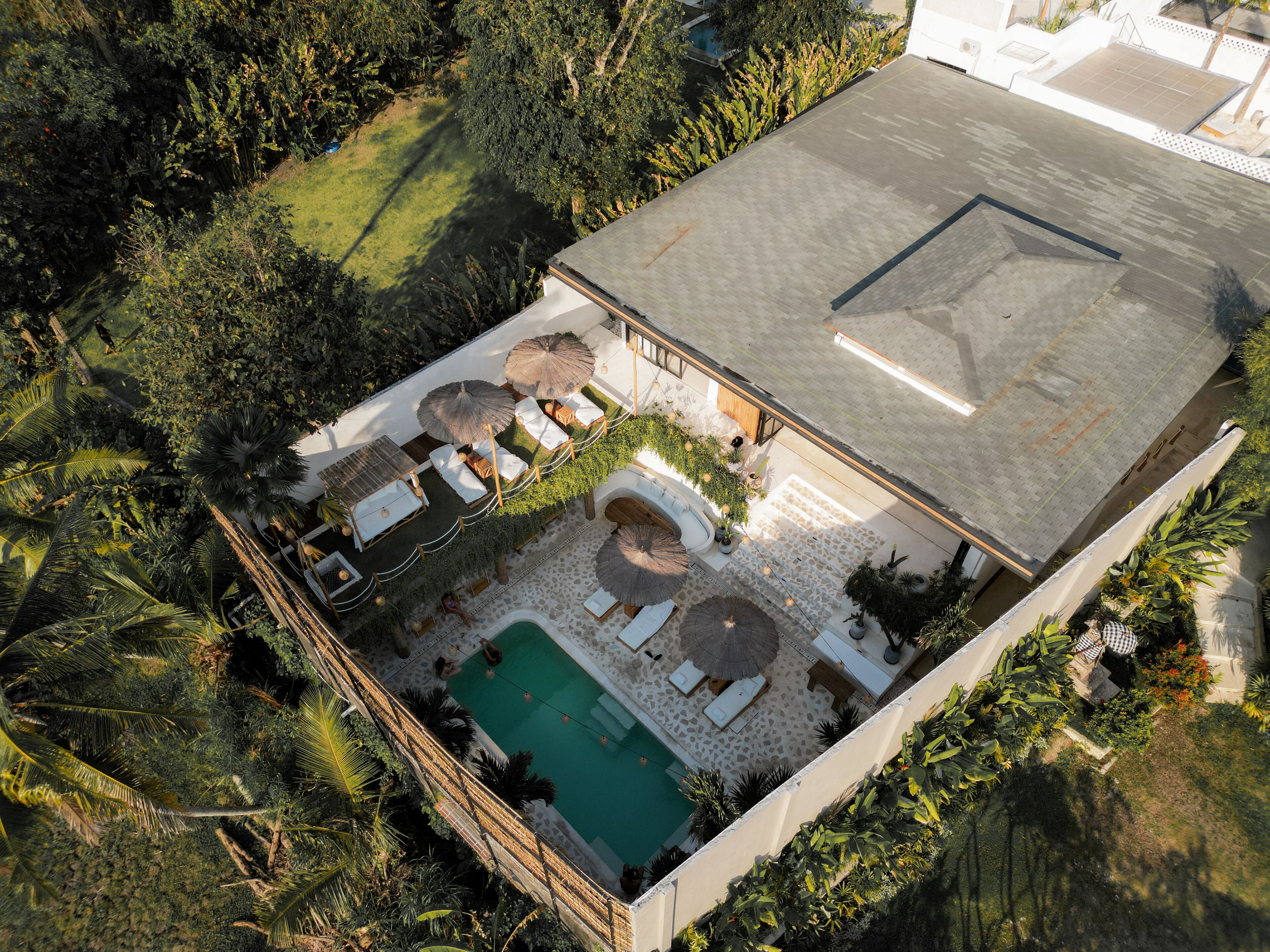 An aerial view of a property with a swimming pool, sun loungers, and a covered patio area surrounded by lush greenery.
