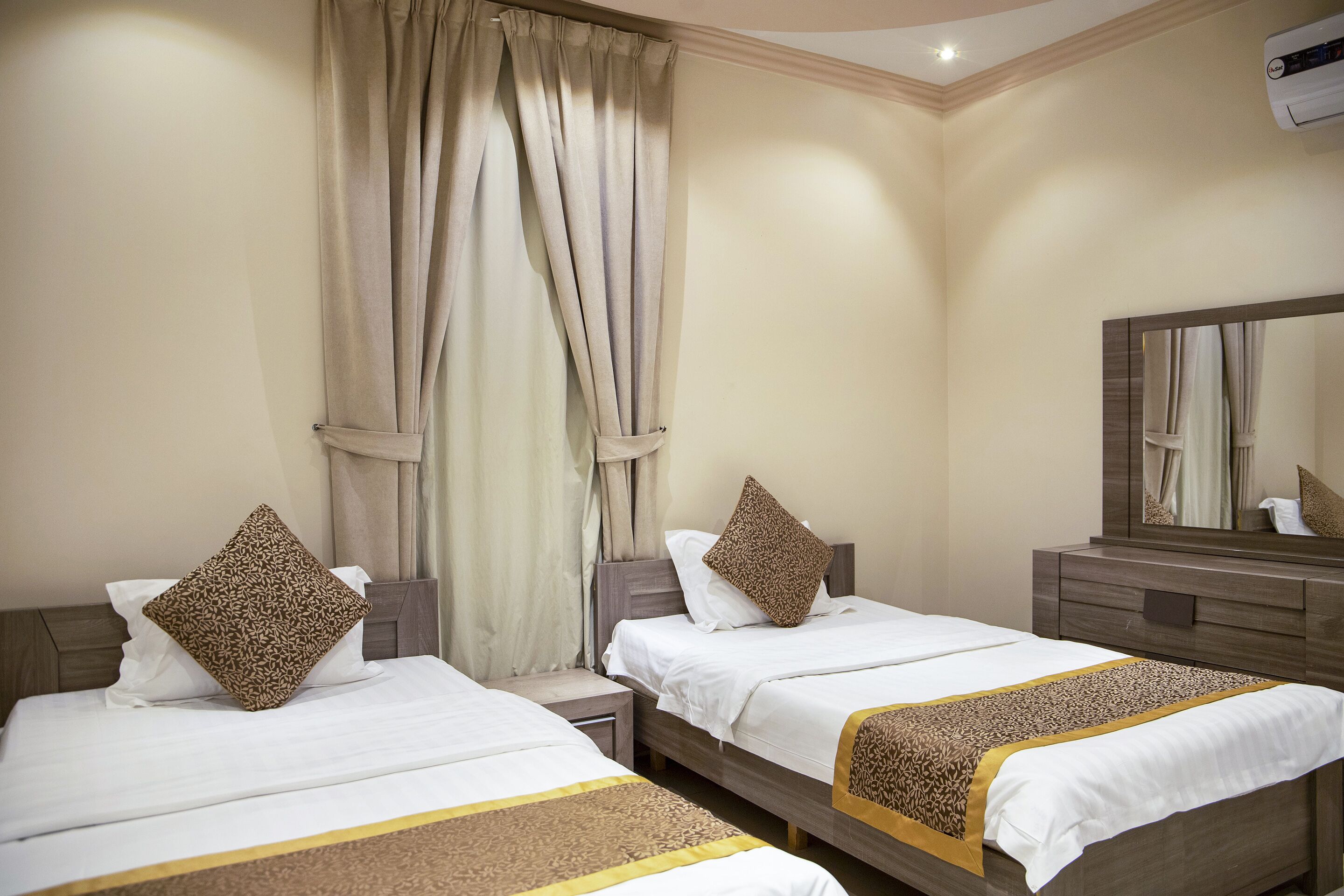 Een hotelkamer met twee bedden, elk met een witte spreidek en een bedgooi. Er hangt een grote spiegel aan de muur en er is een raam met gordijnen.