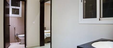 A bathroom with a white sink, a shower, and a toilet. There is a door leading to a bedroom with a bed.