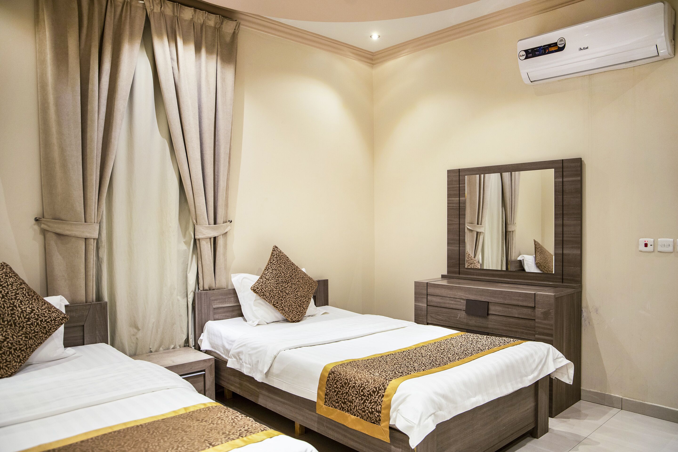 A hotel room with two beds, beige curtains, a wooden nightstand, a mirror, and an air conditioning unit.