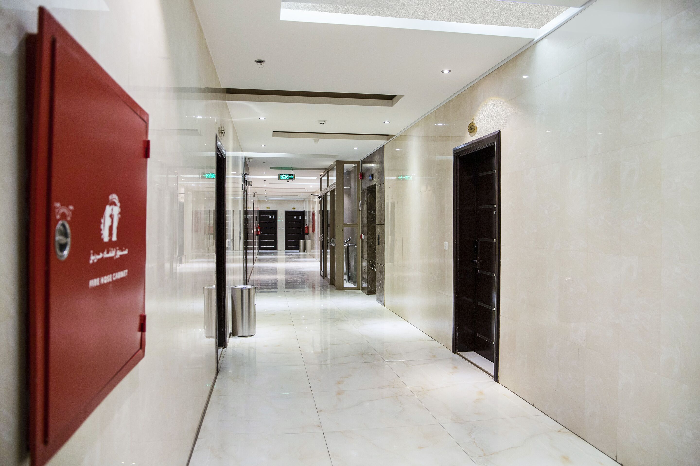 Un pasillo de hotel moderno y limpio, con un armario rojo de seguridad contra incendios a la izquierda, una puerta de ascensor negra a la derecha y un suelo de mármol brillante.